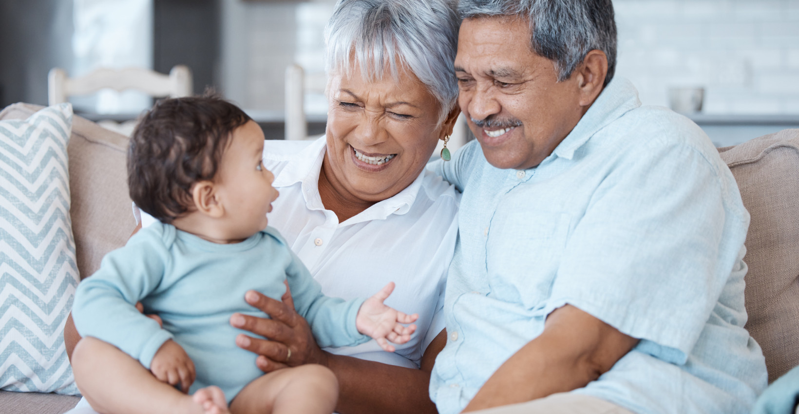 An elderly couple sitting on their couch with their infant grandchild. 