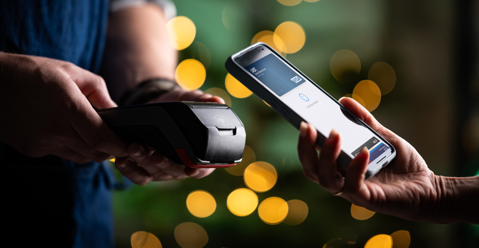 two people stand near each other, one person holding a payment machine, the other holding their phone to the payment machine