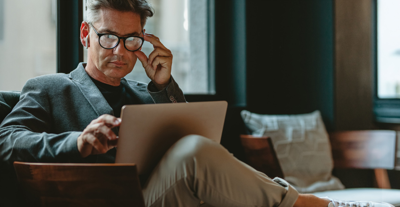 a man sits in a chair with his leg up, adjusting his glasses while looking at a laptop
