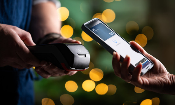two people stand near each other, one person holding a payment machine, the other holding their phone to the payment machine