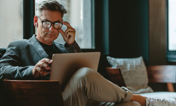 a man sits in a chair with his leg up, adjusting his glasses while looking at a laptop