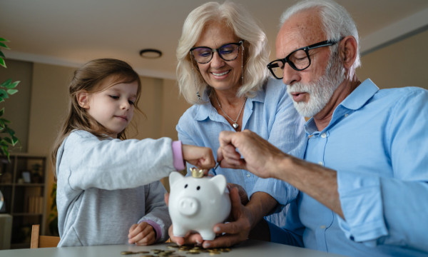 a grandma and grandpa help their granddaughter put coins into a white piggy bank